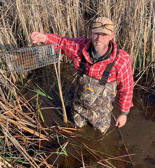 Adam Ahlers standing in water holding cage with beaver inside Adam Ahlers standing in water holding cage with beaver inside