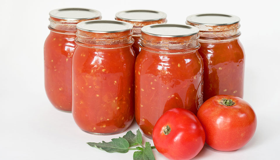 Five jars of canned tomatoes, and two tomatoes sitting on table Five jars of canned tomatoes, and two tomatoes sitting on table