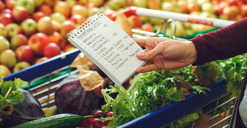 Close-up of woman's hand holding a grocery shopping list, with various vegetables in a basket and in the background Close-up of woman's hand holding a grocery shopping list, with various vegetables in a basket and in the background