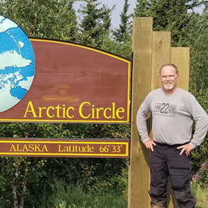 Man with grey shirt standing next to a wooden sign that reads Arctic Circle