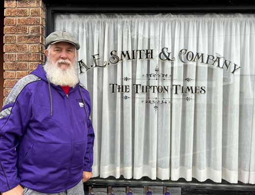 Man with gray beard wearing hat standing in front of business, Tipton Times newspaper