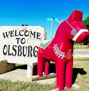 Red Swedish dala horse, painted, in front of Welcome to Olsburg sign